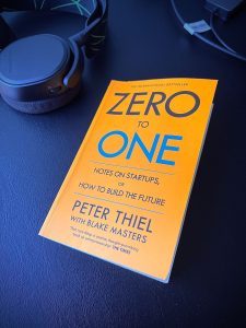 Paperback copy of book “Zero to One” by Peter Thiel and Blake Masters on a black desk next to some headphones.