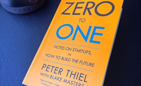 Paperback copy of book “Zero to One” by Peter Thiel and Blake Masters on a black desk next to some headphones.