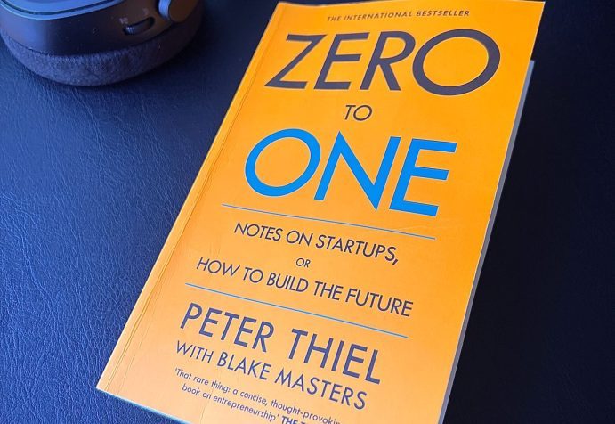 Paperback copy of book “Zero to One” by Peter Thiel and Blake Masters on a black desk next to some headphones.